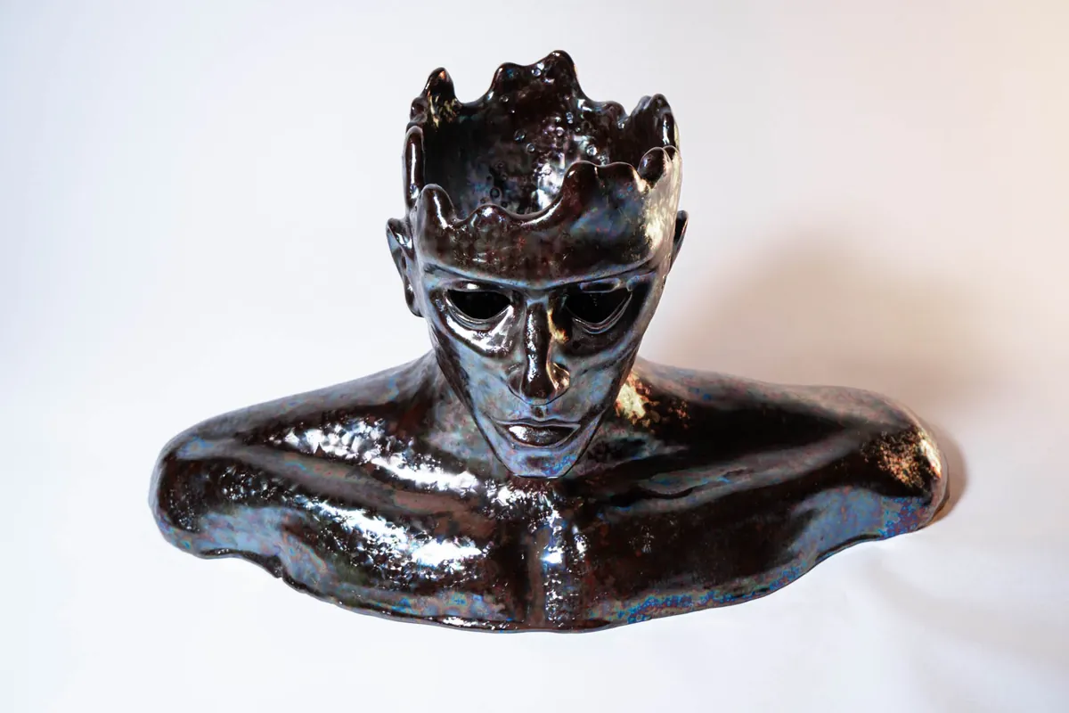 Chrysalis: a glossy ceramic bust with a hollow, crown-like open skull and empty eye sockets, its dark specialty glaze shimmering blue, violet and copper.