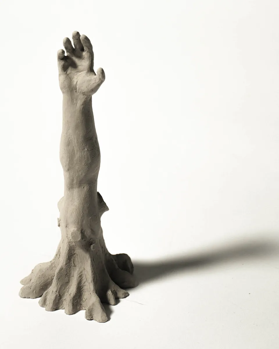 An unfired stoneware arm rising upright from a root-like base, hand open at the top, casting a long shadow on a white surface.