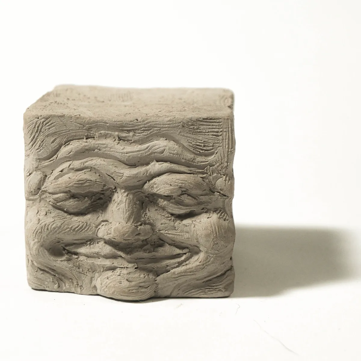 A small stoneware cube with a wrinkled, smiling face sculpted into its front, on a white surface.