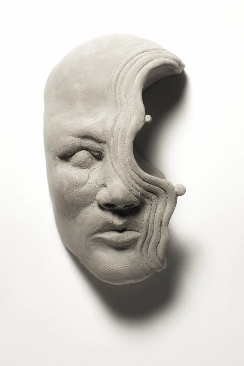 A half-face stoneware wall piece: a calm face with closed eyes whose right side flows away into wavy, crescent-shaped ridges.