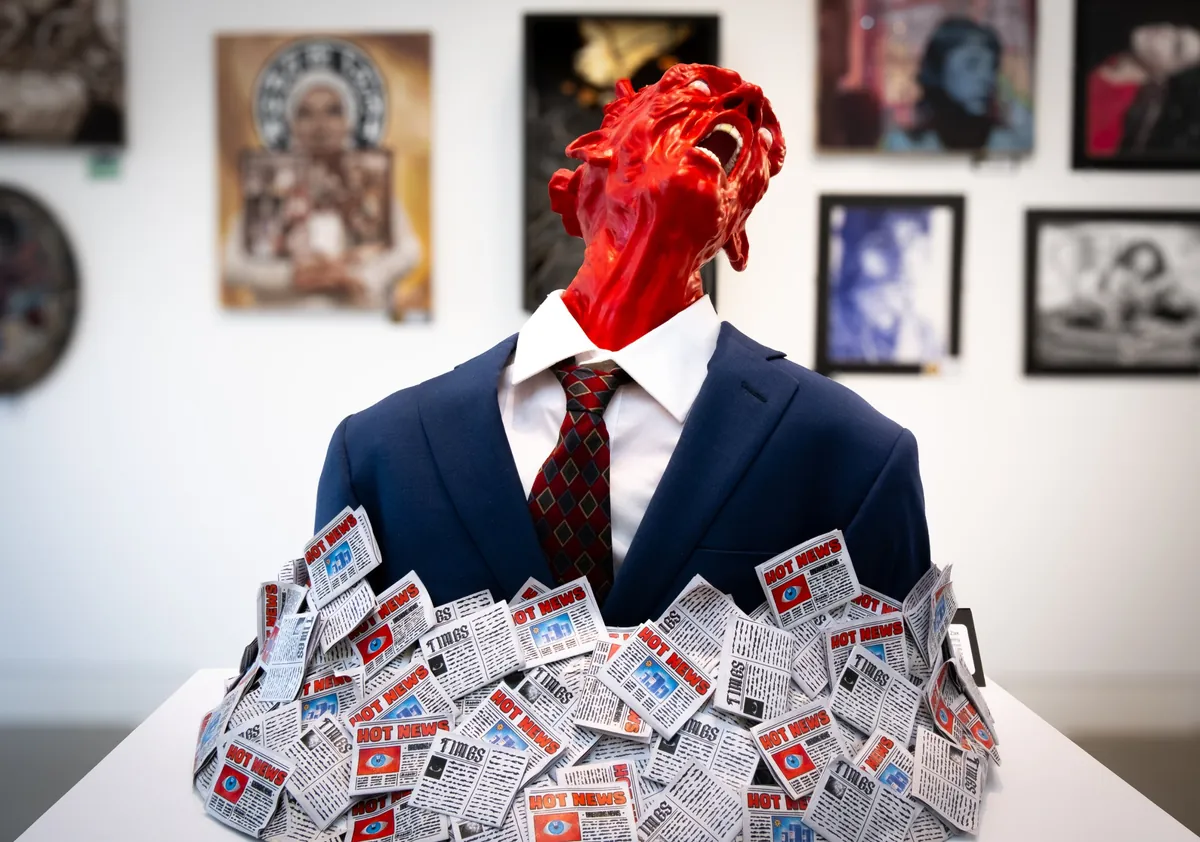 Hots in a gallery: a glossy red ceramic head tipped back mid-scream on a navy-suited bust, buried to the chest in tiny 'Hot News' newspapers.