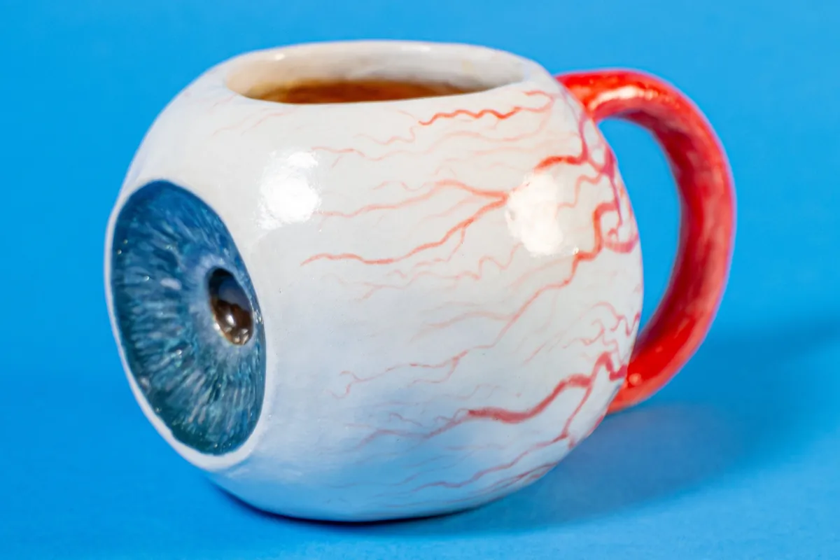 Peep Mug: a round ceramic mug shaped like an eyeball, with a glossy blue iris on the front, hand-painted red veins and a red handle, on a blue backdrop.