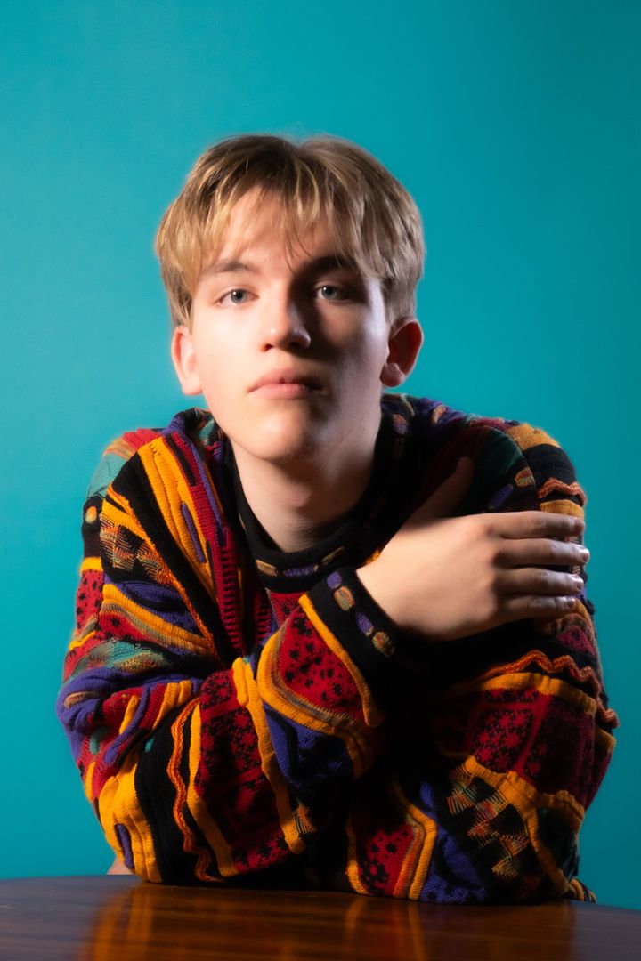 Portrait of Zak Ferenczhalmy against a teal backdrop, leaning on a wooden table in a bright patterned knit sweater.