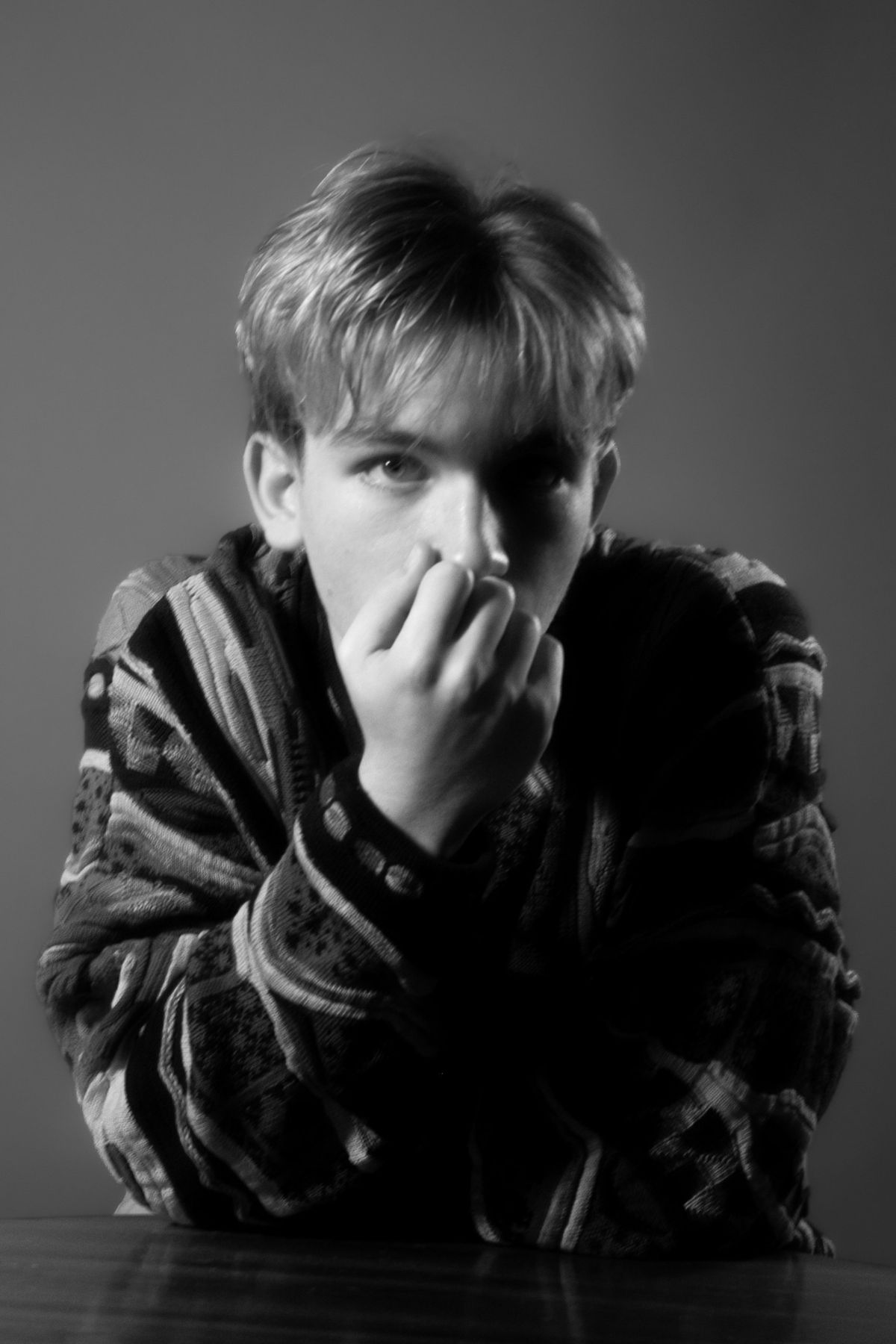 Black-and-white portrait of Zak Ferenczhalmy seated at a table, hand raised to his face, wearing a patterned knit sweater.